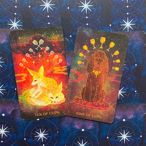 Tarot kortos – dvi atvertos kortos (Ten of Cups su oranžinėmis lapėmis, Nine of Cups su rudu šunimi) ant mėlyno žvaigždėto fono