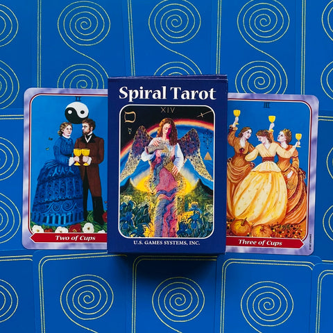 Spiral Tarot kortų rinkinys su mėlyna dėže centre, priekyje matomos Two of Cups ir Three of Cups kortos, pabrėžia pakavimą ir iliustracijas