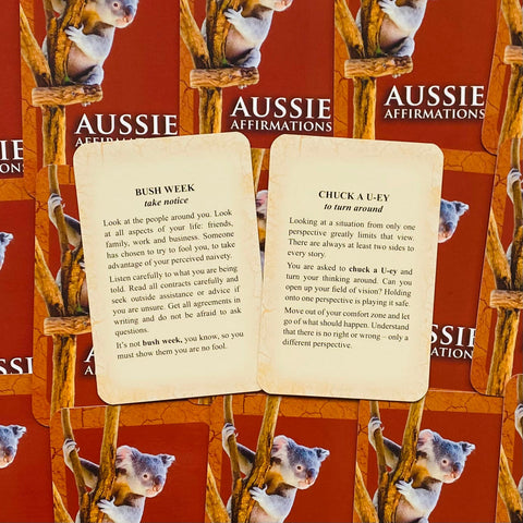 Aussie afirmacijų kortelės, dvi atverstos kreminės kortos su tekstais Bush Week ir Chuck a U-ey prieš raudoną dėžutę su koala