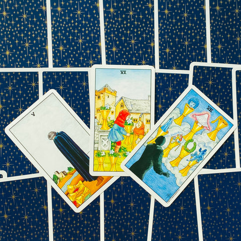 Tarot kortos su mėlynu žvaigždėtu nugarėliu, trys Taurių kortos centre skirtingais kampais, aiškūs piešiniai ir ryškios spalvos