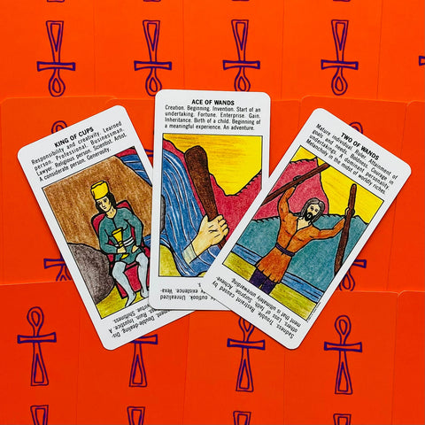 Tarot kortos su ryškiai oranžine nugarėle, trys kortos atvertos (Ace of Wands, Two of Wands, King of Cups) plokščias viršaus vaizdas
