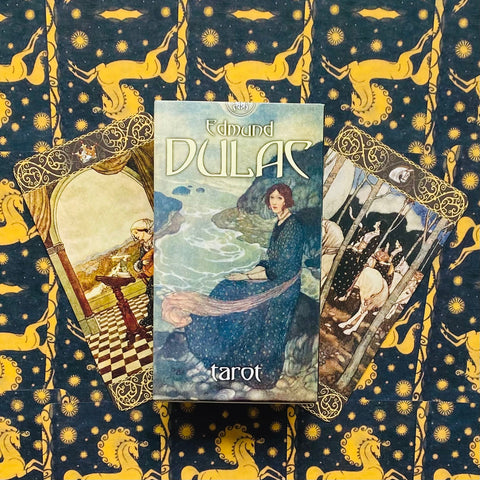 Edmund Dulac tarot kortų rinkinys su mėlyna iliustruota dėžute ir dviem atverstomis kortomis ant geltonai‑mėlyno fono