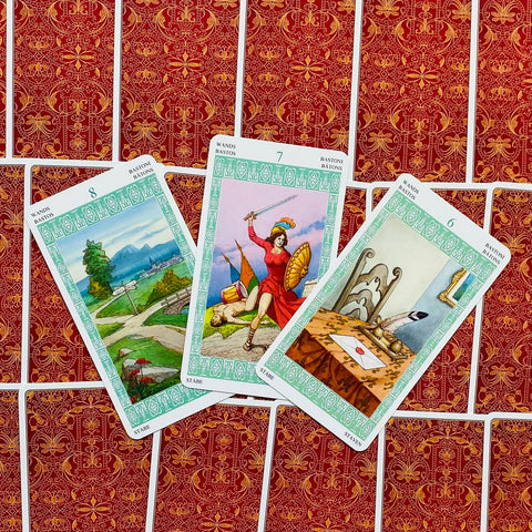 Tarot kortos su žalia‑baltu rėmeliu — trijų lazdų kortų variantai atversti ant raudonų nugarėlių, artimas kampas