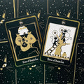 Tarot kortos juodos‑auksinės su katėmis, dvi priekinės kortos — Keturi pentakliai ir Trys pentakliai ant dėklo