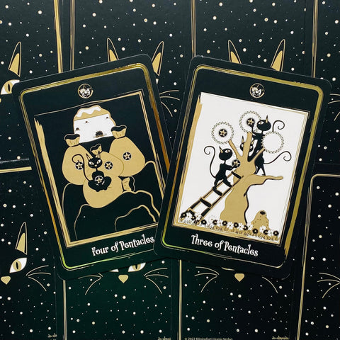 Tarot kortos juodos‑auksinės su katėmis, dvi priekinės kortos — Keturi pentakliai ir Trys pentakliai ant dėklo