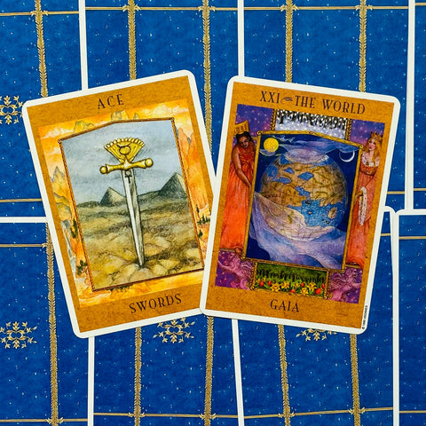 tarot kortos Gaia, atverstos Ace of Swords ir XXI The World kortos ant mėlyno ornamentuoto fono, matomas kortų priekinis dizainas