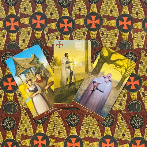 Tarot kortos atverstos trijulė: 2, 3 ir 4 lazdų kortos ant raudono‑geltono ornamentuoto fono, parodytos priekinės pusės