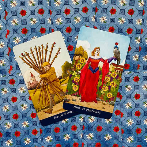 Tarot kortos Rider–Waite stiliaus, dvi kortos (Ten of Wands, Nine of Pentacles) ant mėlyno gėlėto fono, pasuktos kampu