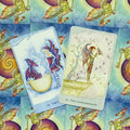 Tarot kortos su pastelinių mėlynų ir žalsvų atspalvių drakonų bei sparnuotų figūrų iliustracijomis, dvi skirtingos kortos parodytos iš priekio, skirtingos variacijos parduotuvės galerijoje