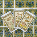 Tarot kortos su baltu rėmeliu, fanuotos trys Lazdų kortos (5–7) centre, padėtos ant mėlyno‑žalsvo ornamentuoto nugarėlių fono