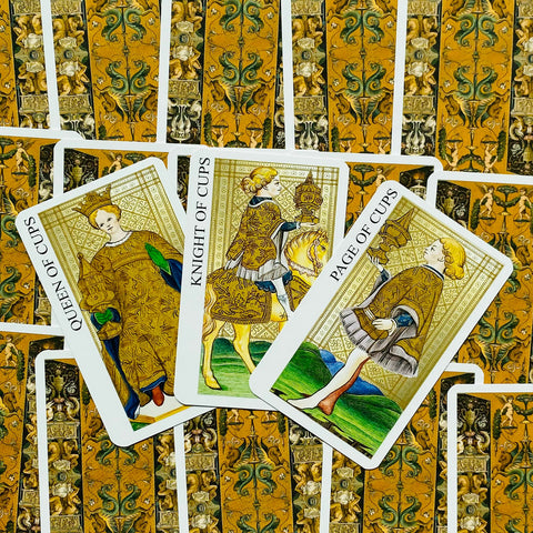 Tarot kortos su aukso rašto nugarėlėmis ir atvertomis Taurų kortomis — Karalienė, Riteris ir Puslapis matomi iš viršaus