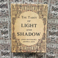 Tarot kortos Light and Shadow – dėžutė su smėlio spalvos viršeliu ir sudėtingais ornamentais, matoma iš priekio ant raštuoto kortų fono