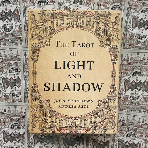 Tarot kortos Light and Shadow – dėžutė su smėlio spalvos viršeliu ir sudėtingais ornamentais, matoma iš priekio ant raštuoto kortų fono