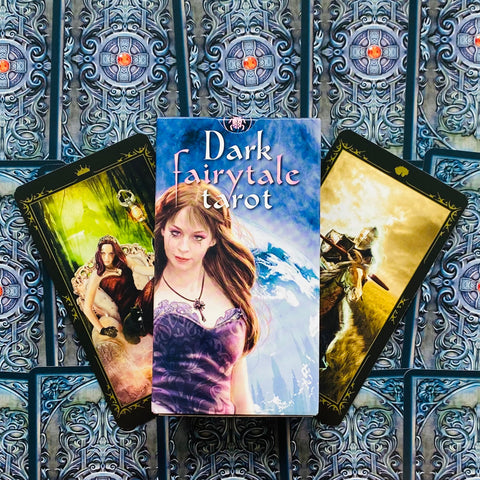 Dark Fairytale Tarot kortos dėžutė priekyje su dviem atvertais kortomis, violetinė dėžutė ir gotikinis raštuotas fonas, aiškus produkto rodymas pirkėjui