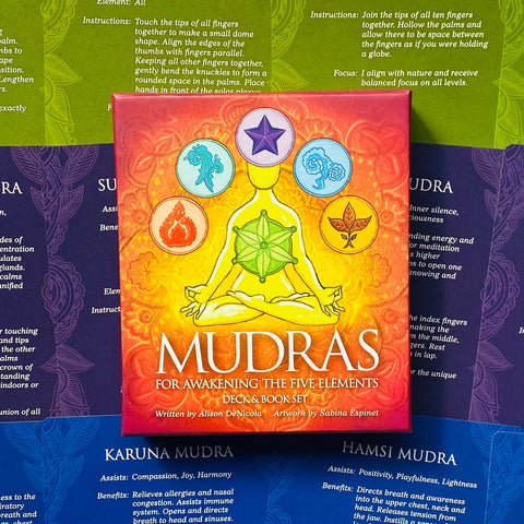 Mudras kortų rinkinys ryškiai oranžinė dėžutė su meditacijos simboliais centre, padėta ant mėlynų ir žalių vadovo puslapių