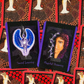 Orakulo kortos: dvi atviros kortos – mėlyna sparnuota Sacred Learning ir portretinė Magickal Dreaming su purpuriniais kraštais ant raudono taškuoto nugarėlio