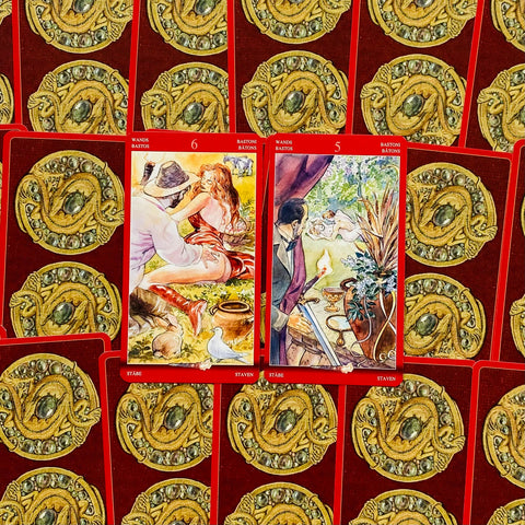 Tarot kortos su raudonomis nugarėlėmis, dvi atviros kortos (6 ir 5 lazdų) su spalvingomis iliustracijomis ir auksiniu ornamentu fonu