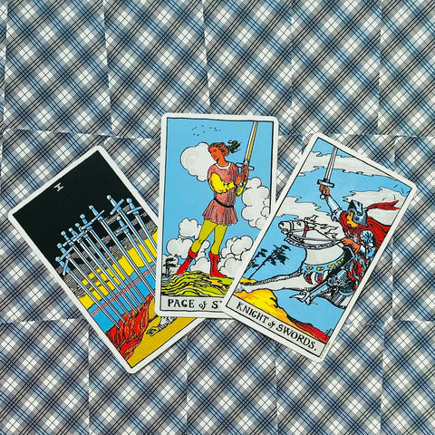 tarot kortos Rider–Waite trijų kortų išklotinė: centre Page of Swords, dešinėje Knight of Swords, šviesiai mėlynas raštuotas fonas