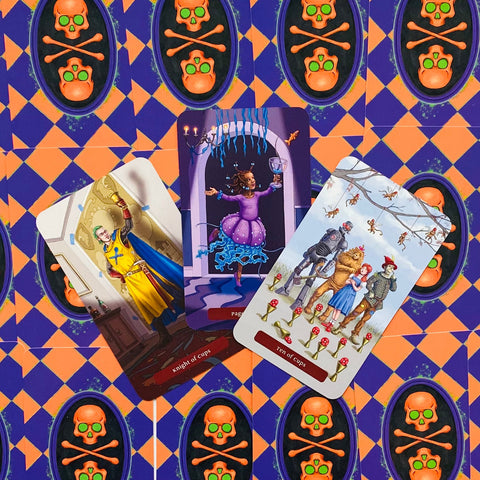 Tarot kortos iliustruotas rinkinys su purpurine‑oranžine kaukolės nugarėle, trys atverstos kortos: riteris, šokanti dama ir dešimt taurų šeimos scena