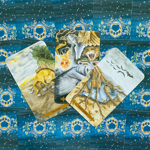 Tarot kortos su gyvūnų iliustracijomis, trys atverstos kortos centre – šaknys, žaibas ir ugnis, tamsiai mėlynomis nugarėlėmis