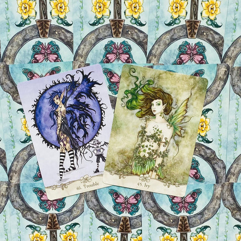tarot kortos su fėjų iliustracijomis, dvi atverstos kortos — "Trouble" (mėlyna–violetinė) ir "Ivy" (žalia) ant ornamentuoto fono