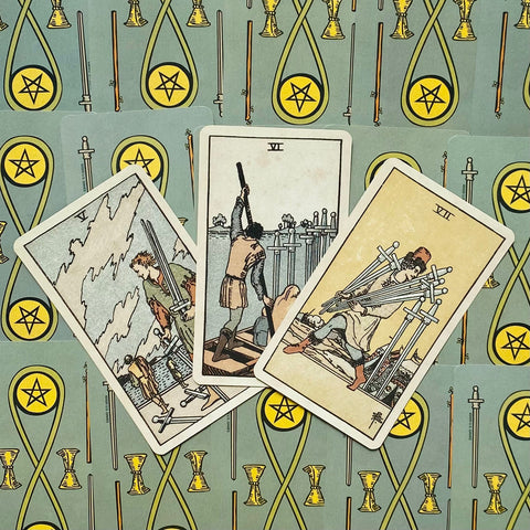 Tarot kortos trys Kardų kortos (V–VII) išdėstytos ant žalsvo pentaklių rašto fono, užfiksuota iš arti kampu