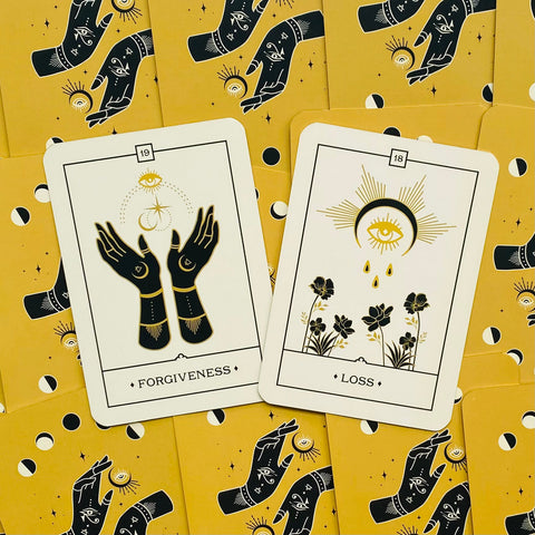 Tarot kortos, atviros baltos kortos „Forgiveness“ ir „Loss“ su juodai auksinėmis iliustracijomis ant geltonų nugarėlių, išdėstytos plokščiai prekybinėje nuotraukoje