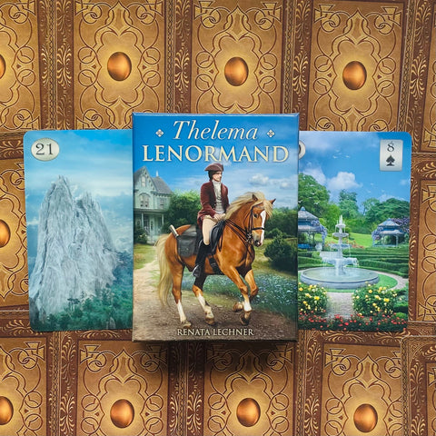Lenormand kortų rinkinys Thelema, mėlyna dėžutė su raiteliu (Renata Lechner) ir dvi atviros kortos šonuose, ornamentuotu fonu
