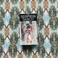 Serpieri tarot kortų rinkinys ant ornamentuoto fono, frontali dėžutė su moters iliustracija ir viršaus logotipu matoma