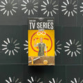 TV Series Tarot dėžutės priekinis vaizdas: geltona pakuotė su figūra laikančia kortą, centroje ant juodo raštuoto fono