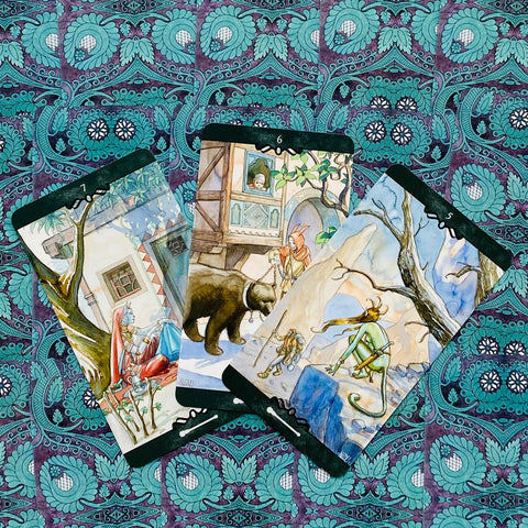 Tarot kortos su akvarelinėmis iliustracijomis — trys kortos vėduoklėje ant turkio raštuoto audinio, rodoma kortų priekinė pusė
