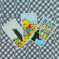 Tarot kortos Rider–Waite, trys taurės (V, VI, VII) iš 78 kortų komplekto, išdėstytos ant mėlyno raštuoto audinio, matomos iš viršaus