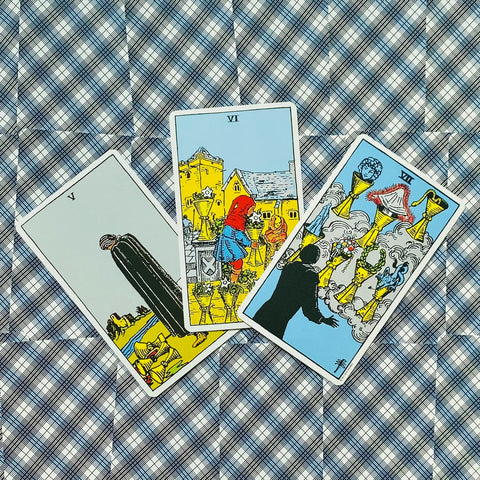Tarot kortos Rider–Waite, trys taurės (V, VI, VII) iš 78 kortų komplekto, išdėstytos ant mėlyno raštuoto audinio, matomos iš viršaus