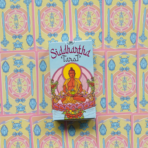 Siddhartha tarot kortos baltai-mėlyname dėkle su sėdinčio Budos iliustracija, centre ant pastelinio kortų fono