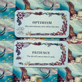Inspiracinės kortelės su jūros motyvais, pastelinė mėlyna fone, dvi kortelės „Optimism“ ir „Patience“ išdėstytos viena virš kitos