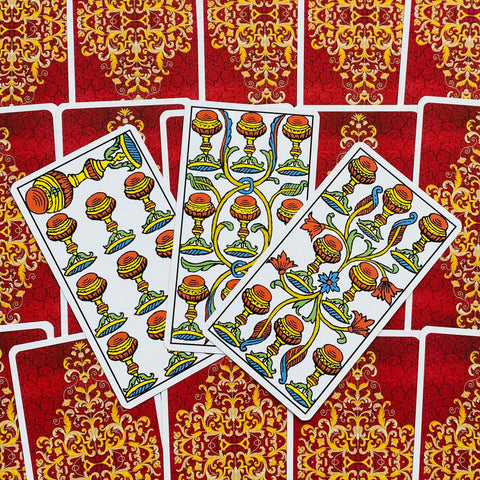 Tarot kortos su raudonomis nugarėlėmis ir auksiniu ornamentu, trys atverstos taurės kortos fane su ryškiomis spalvotomis detalėmis