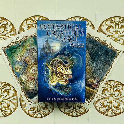 Fantastical Creatures Tarot kortų rinkinys su mėlynu dėklu ir trimis iliustruotomis kortomis, išskleistomis ant aukso ornamentų fono