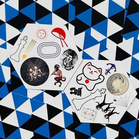 lipdukai — dvi šešiakampės lakšteliai su įvairiais dizainais (maneki‑neko, šachų karalius, skeletas, moneta) ant mėlyno trikampio fono kampiniu žiūrėjimu