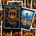 Aboriginal Spirit Oracle kortų rinkinys (Mel Brown) juodame aukso dėkle, išrikiuotas fone, šalia matoma Manifestation korta