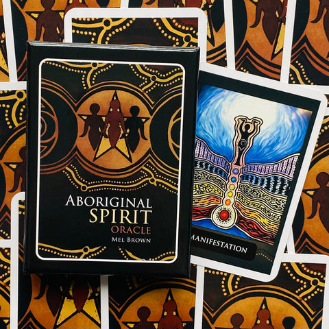 Aboriginal Spirit Oracle kortų rinkinys (Mel Brown) juodame aukso dėkle, išrikiuotas fone, šalia matoma Manifestation korta