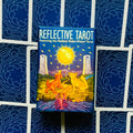 Tarot kortos Reflective Tarot, Radiant Rider‑Waite leidimas mėlyname dėkle, priekinis kampas prieš tamsiai mėlyną modeliuotą kortų nugarėlių foną