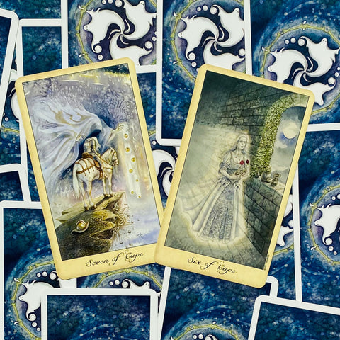 Tarot kortos su mėlynomis nugarėlėmis ir dviem auksuotais, iliustruotais korteliais (Septynios taurės, Šešios taurės) ant išskleistos fono kompozicijos