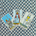 Tarot kortos Rider–Waite, trys kortos: Kardų karalius centre, Kardų karalienė kairėje ir Puodelių tūzas ant languoto audinio