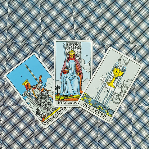 Tarot kortos Rider–Waite, trys kortos: Kardų karalius centre, Kardų karalienė kairėje ir Puodelių tūzas ant languoto audinio