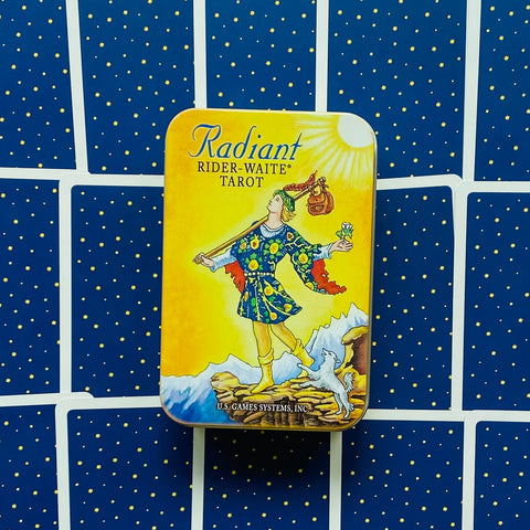 Tarot kortos Radiant Rider‑Waite metalinėje geltonoje dėžutėje, centre ant išskleistų mėlynų kortų nugarėlių su mažomis žvaigždėmis
