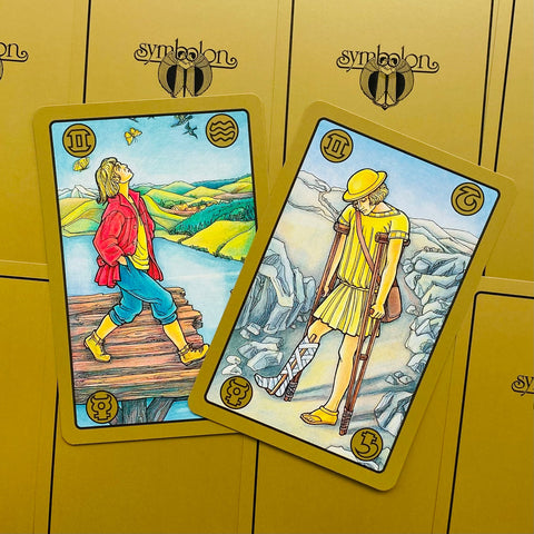 Tarot kortos Symbolon su aukso rėmeliais — dvi atverstos kortos (klajotojas ir ramentinis) ant geltono fono, demonstruoja skirtingus iliustracijų variantus