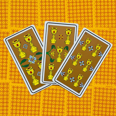 tarot kortos su taurėmis — trys atviros rudos kortos su auksinėmis taurėmis, išdėstytos iš viršaus ant ryškaus geltono raštuoto fono