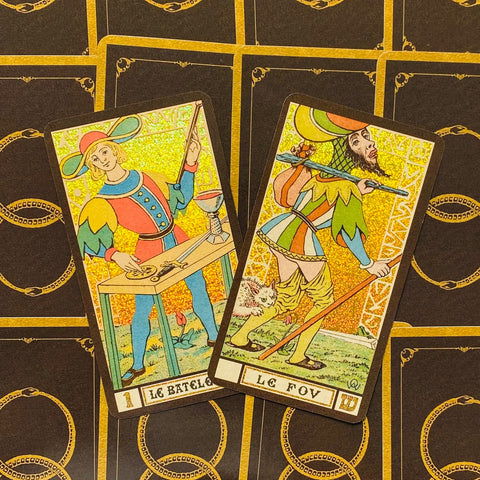 Tarot kortos su auksiniu blizgiu fonu, dvi atverstos kortos (Le Bateleur ir Le Fou) rodomos iš viršaus