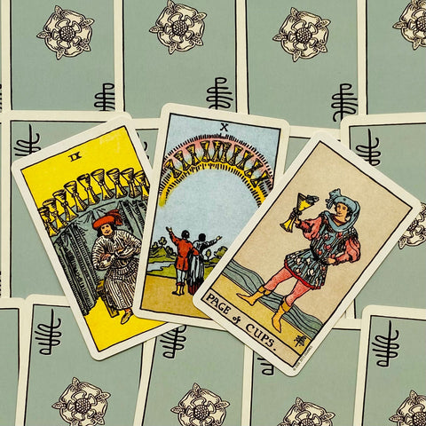Tarot kortos Rider–Waite komplektas — trys atverstos puodelių kortos (IX, X, Page), mėlyna nugarėlė, artimas plokščias kampas