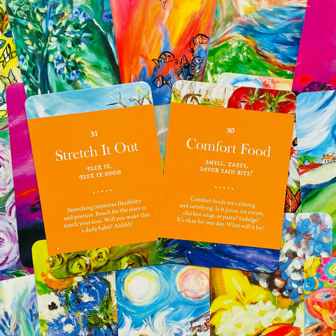 Kortos su oranžiniais užrašais „Stretch It Out“ ir „Comfort Food“, padėtos ant spalvingų tapybos nugarėlių, skirtos kasdieniams įkvėpimams